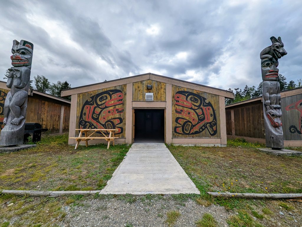 A wooden building with sylized paintings of red and black whales on the walls and two carved wooden totems flanking the building with an overcast sky above.
