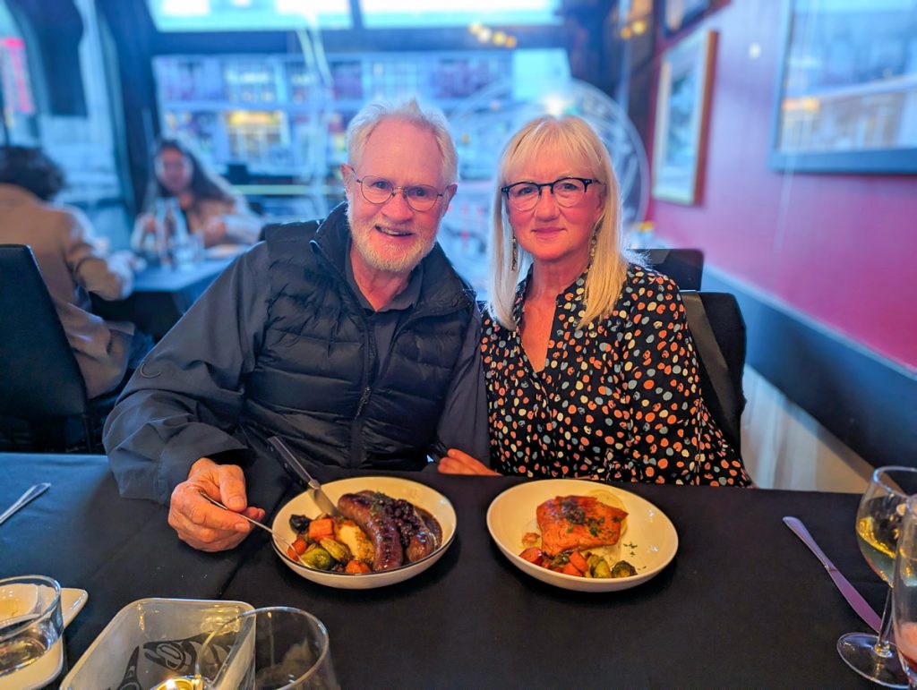 A man and woman both with white hair and glasses smiling at a restaurant table with bowls of yummy food in front of them.