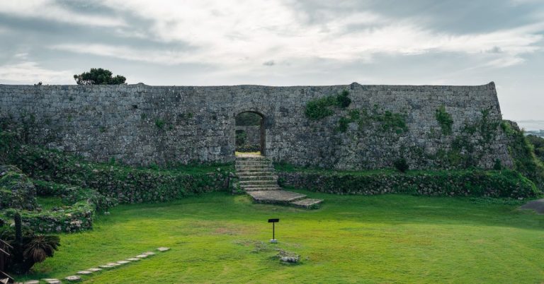 An old stone castle wall with very green grass and a small signpost in the foreground with gray sky in the background.