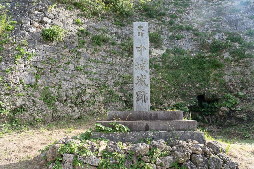 A stone pillar with Japanese text cared into it sitting on more stone with a stone wall behind it.