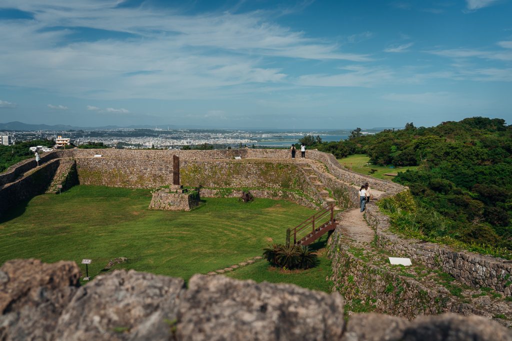 People walking on stone walls surrounding a green field with a city in the distance under blue skies.