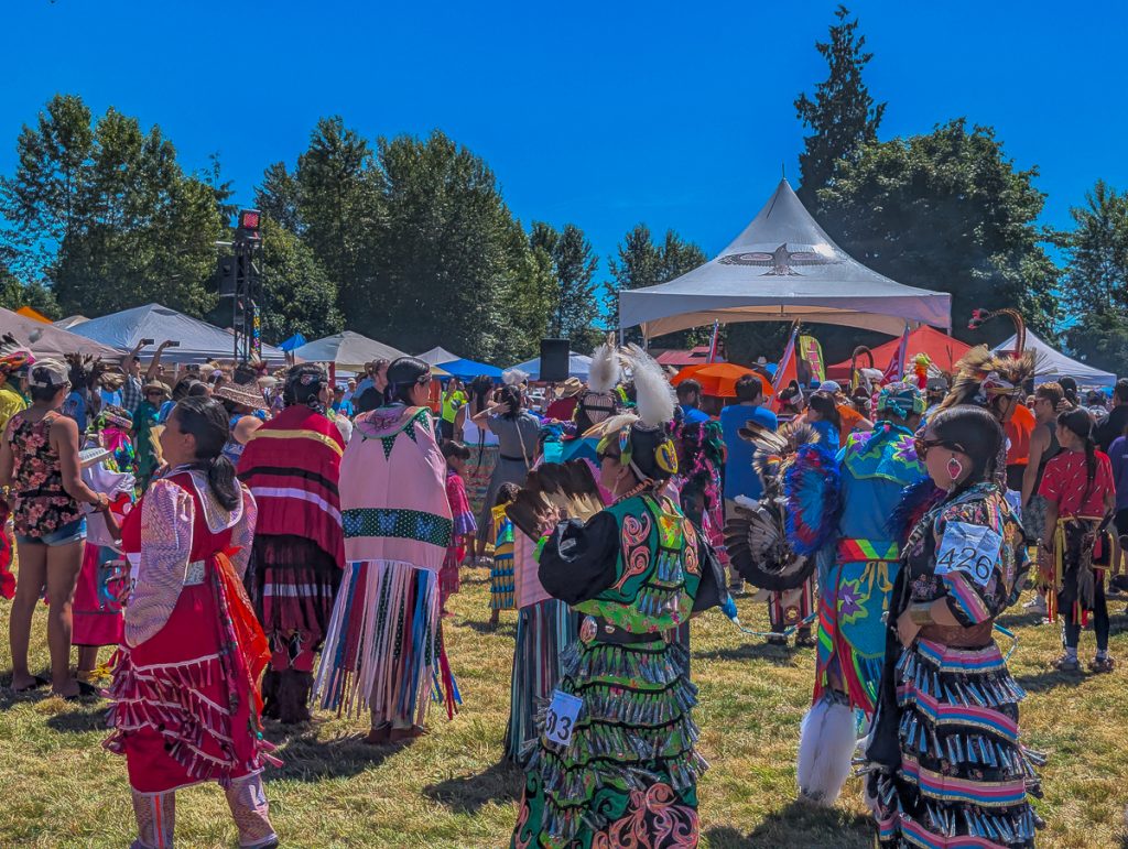 A field with tents and lights with lots of Canadian First Nations people in traditional colorful clothing on a sunny day.