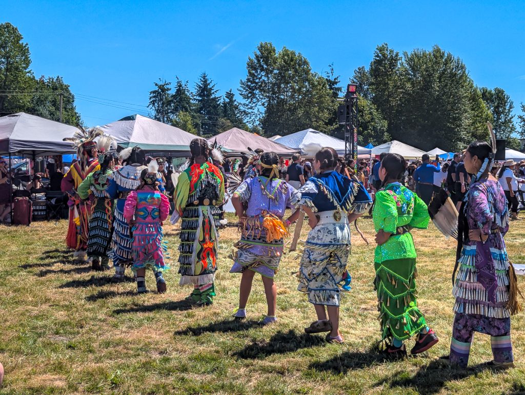 Young Canadian First Nations women in colorful, traditional clothing dancing in a line n a field with tents in the background and blue sky above.