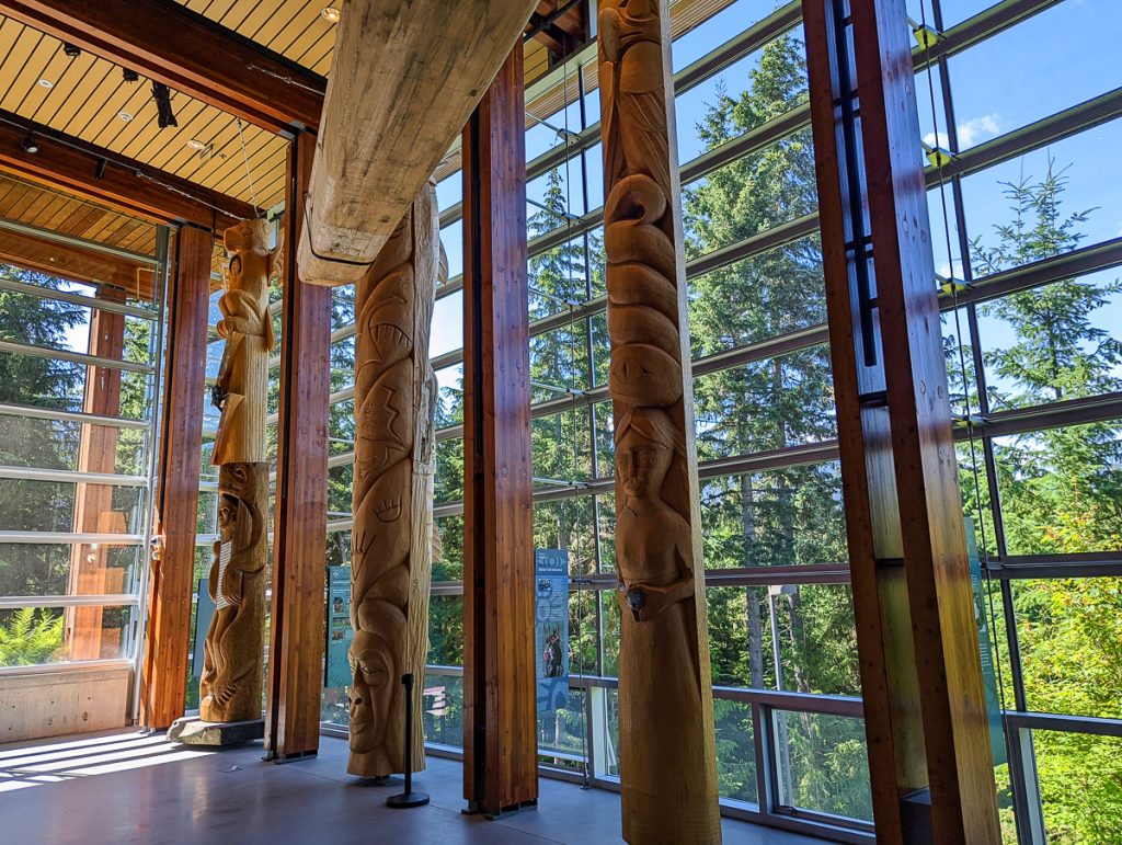 Carved wooden totem poles inside a tall room with floor to ceiling windows looking out on pine trees, blue sky and sunshine.