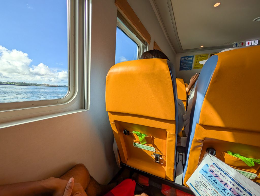Bright orange seats inside a ferry with blue water, green land and blue sky out the window.