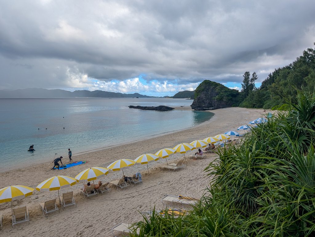 A beach lined with lounge chairs and yellow and white striped umbrellas with low lying clouds above and greenery behind the sand.