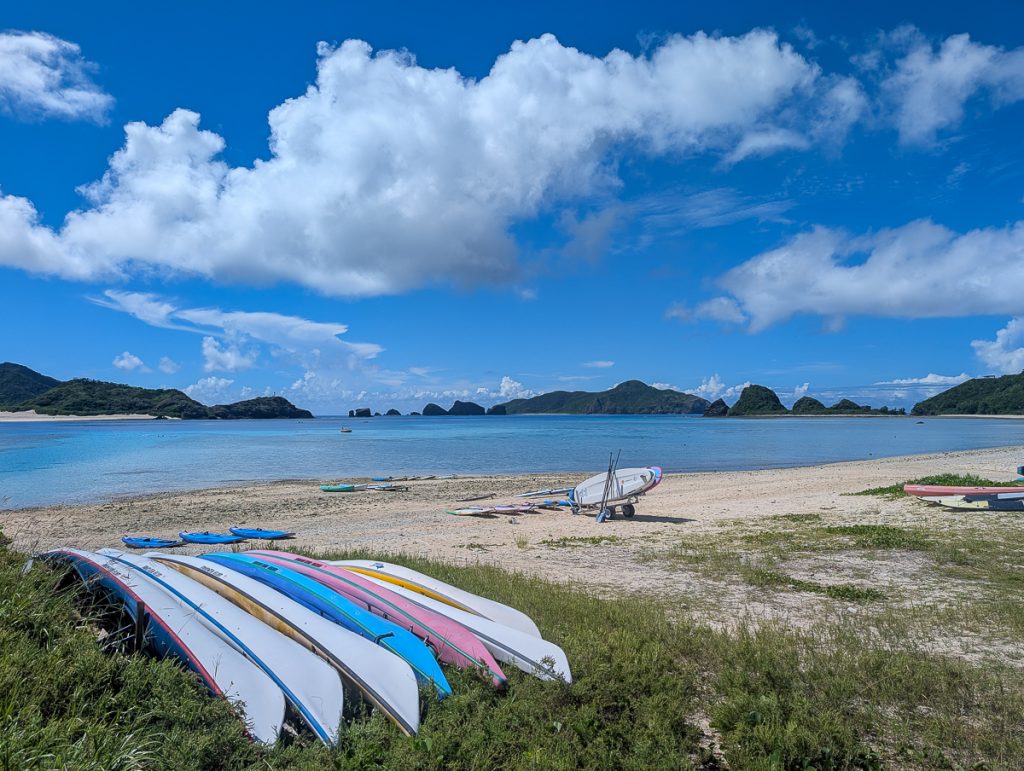 A beach with multiple kayaks and SUP on the sand and shrubbery, with islands in the distance and a blue sky with fluffy white clouds above.