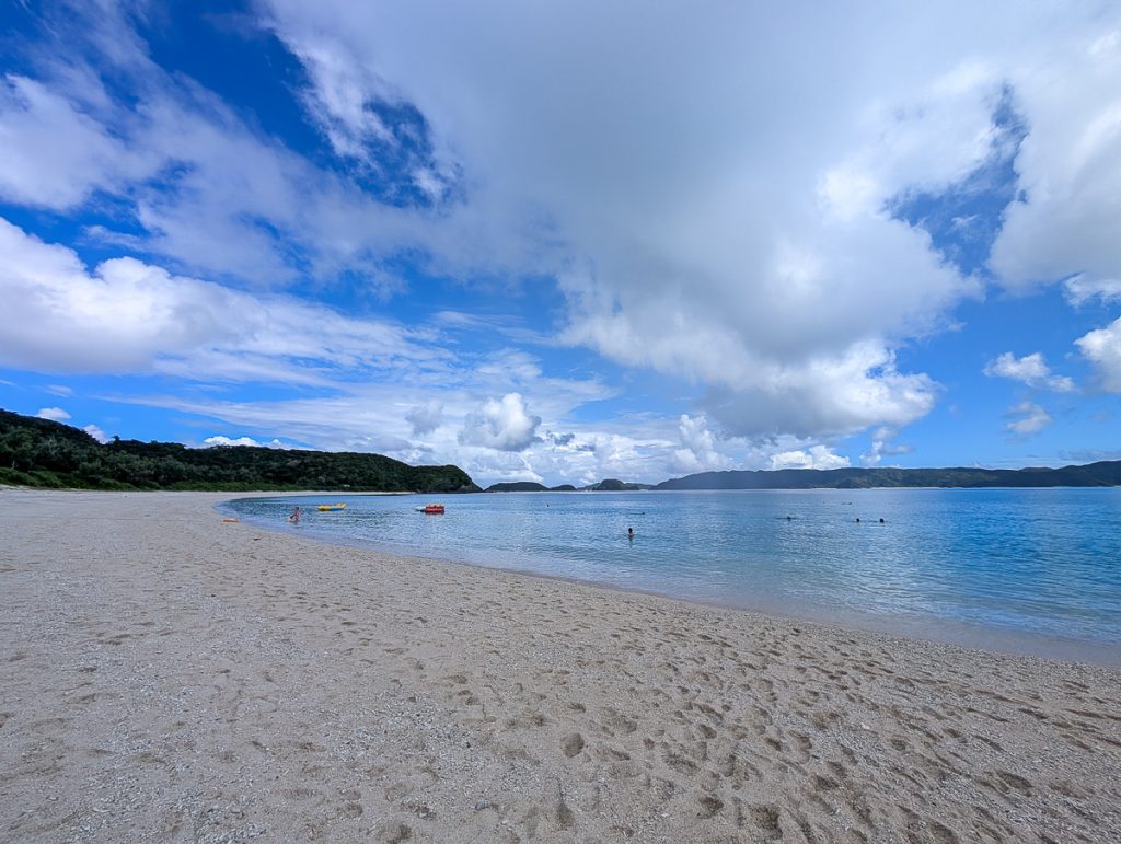 A sandy beach backed by vegetation with a few people kayaking and snorkeling in the waters under blue sky and white clouds.