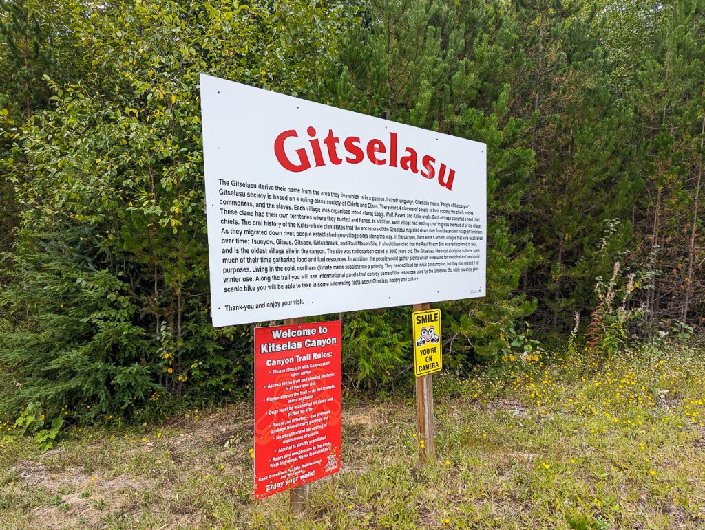 A large white sign with information about the Gitselasu people, with a smaller red sign below it with canyon trail rules for Kitselas Canyon and a small yellow sign saying "Smile you're on camera" with green bushes behind.
