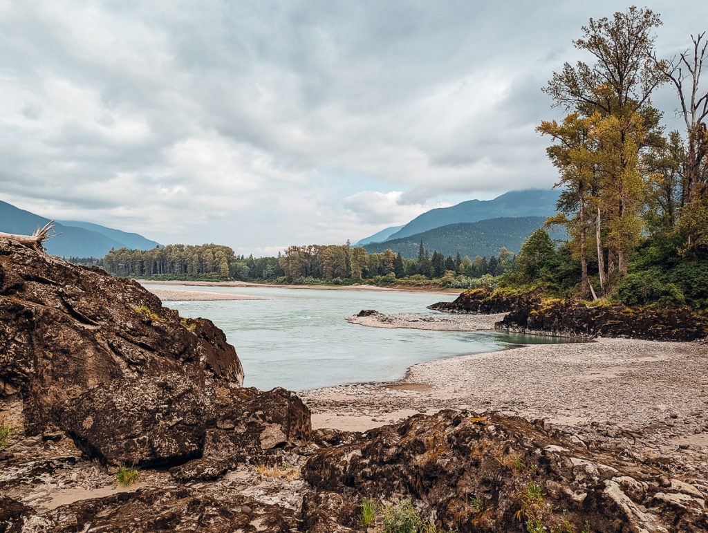 A landscape showing rocks next to a river with trees and mountains in the background and an overcast sky above.