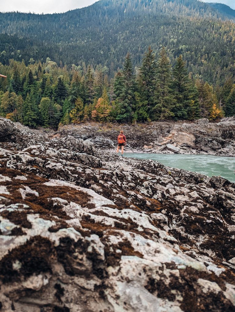 Rocks covered in lichen in the foreground with a person looking tiny next to a river in the background, with pine trees behind.