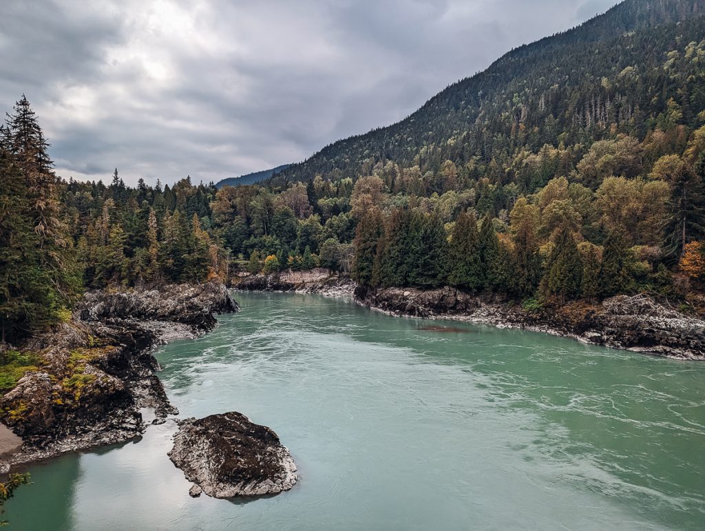 A pale blue-green river with rocks, pine trees and a hill on the banks.