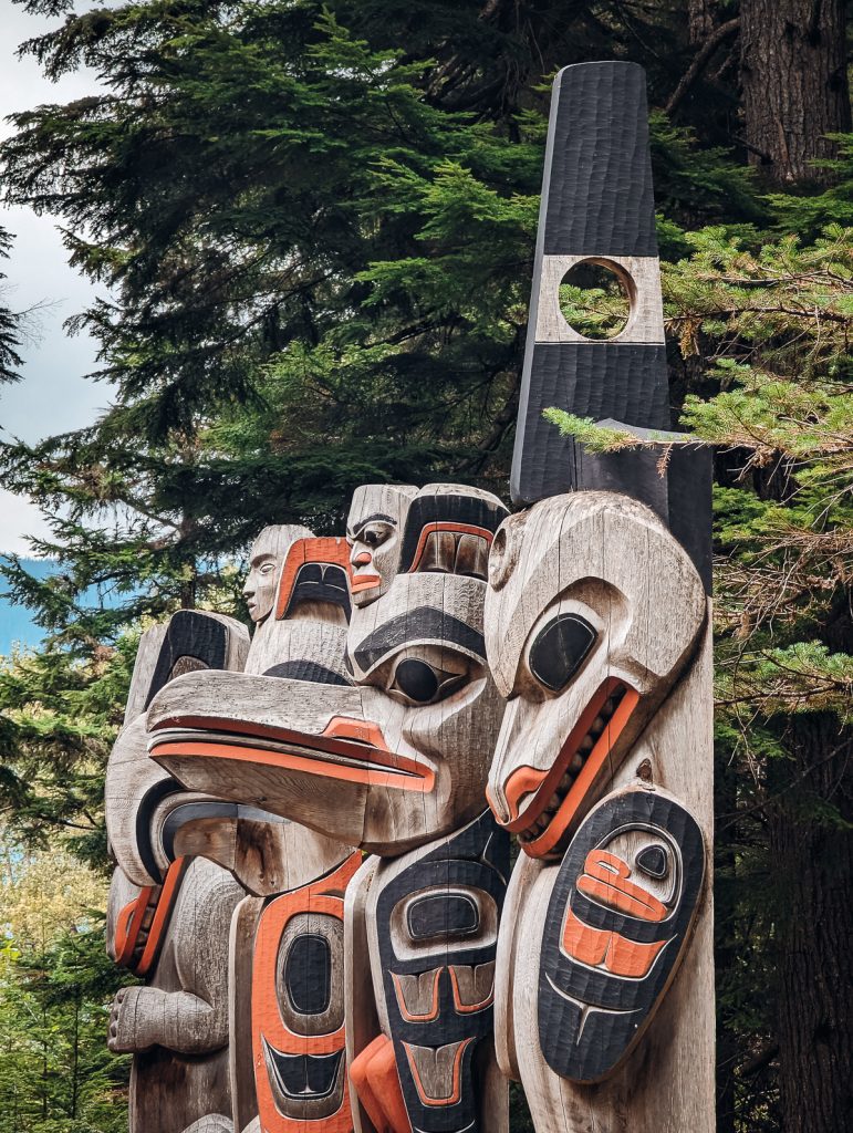 A close up of four carved totem poles seen from the side, with green pine trees behind.