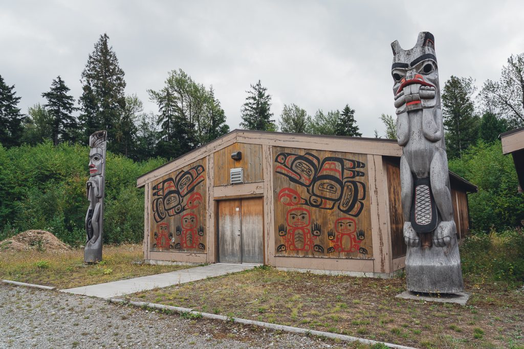 A small wooden building with stylized ravens and people painted on the front in black and red, with two carved totem poles flanking it on either side.