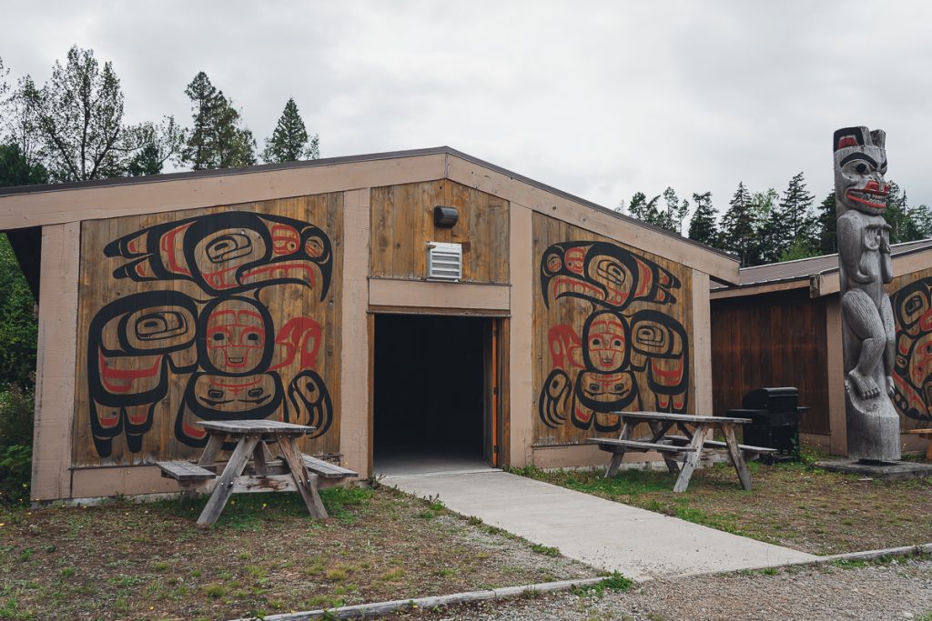 A small wooden building with stylized eagles painted in red and black on the walls, picnic benches sitting outside and a carved totem pole.
