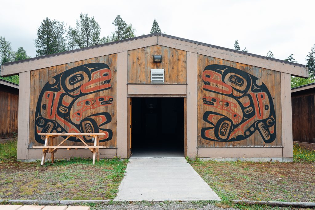 A small wooden building with an open door leading to darkness and stylized orcas painted in black and white on the exterior walls, with a picnic table in front of the building.