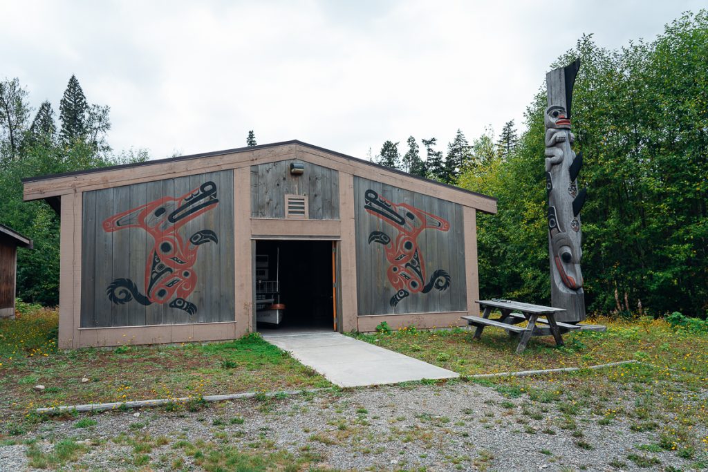 A small wooden building with stylized wolves painted on the walls in black and red, a picnic bench and a totem pole outside.