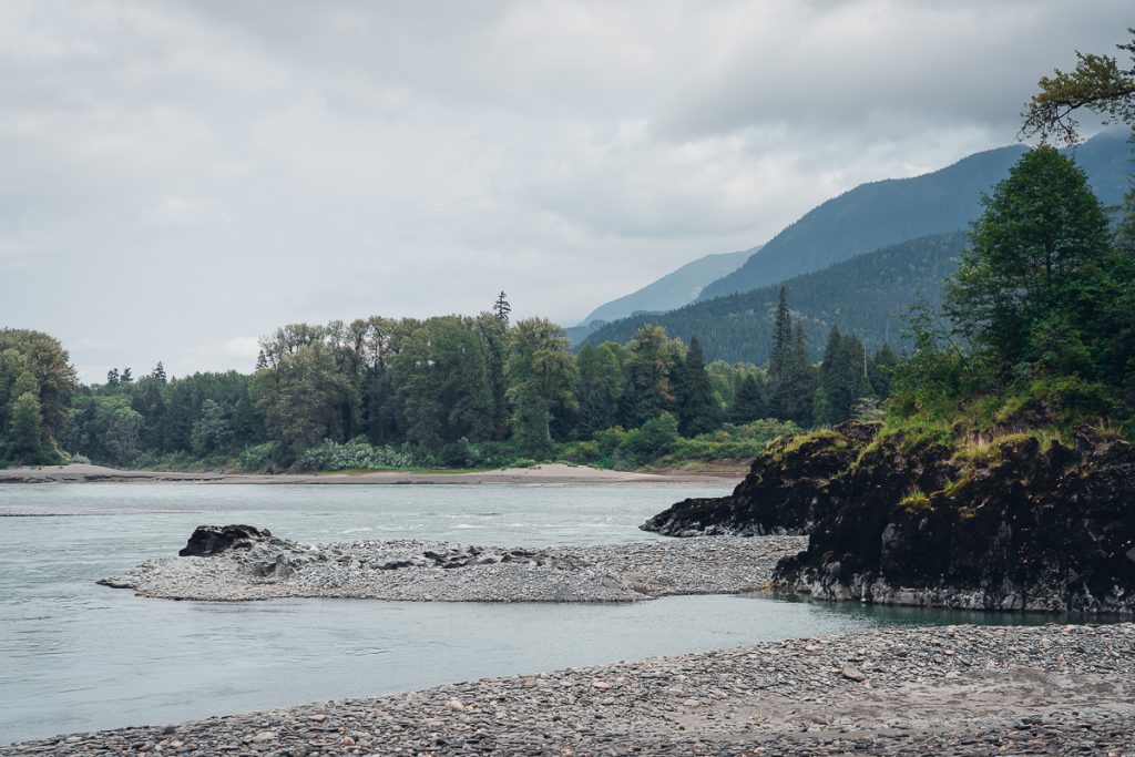 The rocky shore of a calm river with pine trees, mountains and cloudy sky in the background.