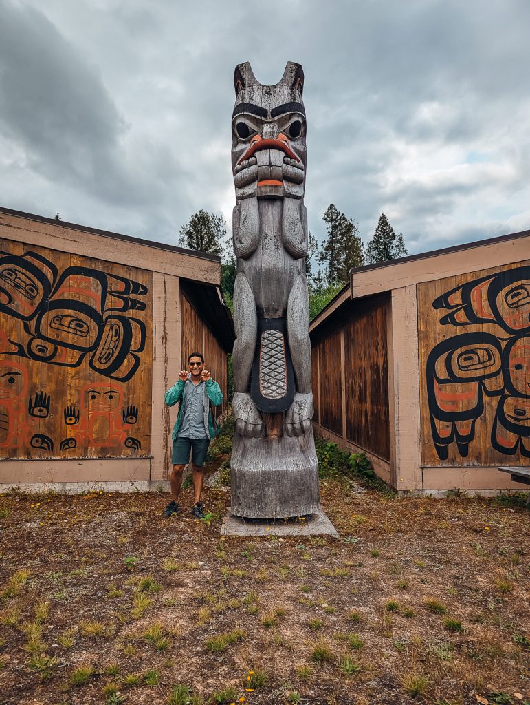 A man standing next to a carved totem pole imitating the figures pose, with two wooden buildings on either side with painted designs on the walls.