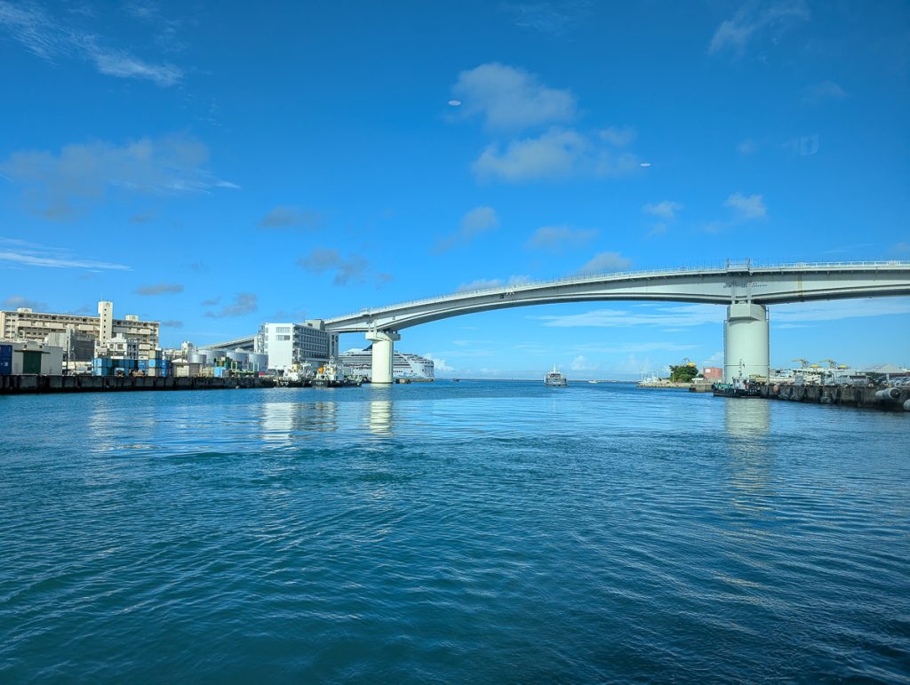 A tall bridge over water with buildings and boats on either side as well as a ferry sailing out towards a cruise ship.