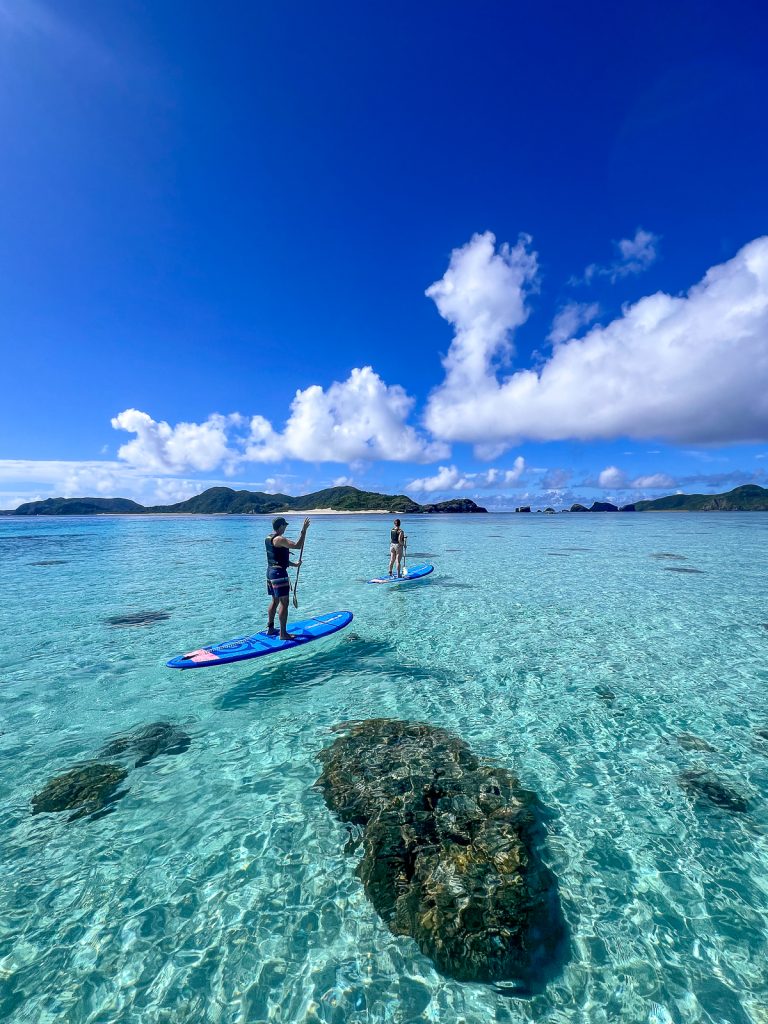 Two people paddling on SUPs across crystal clear waters with blue skies and fluffy white clouds above.