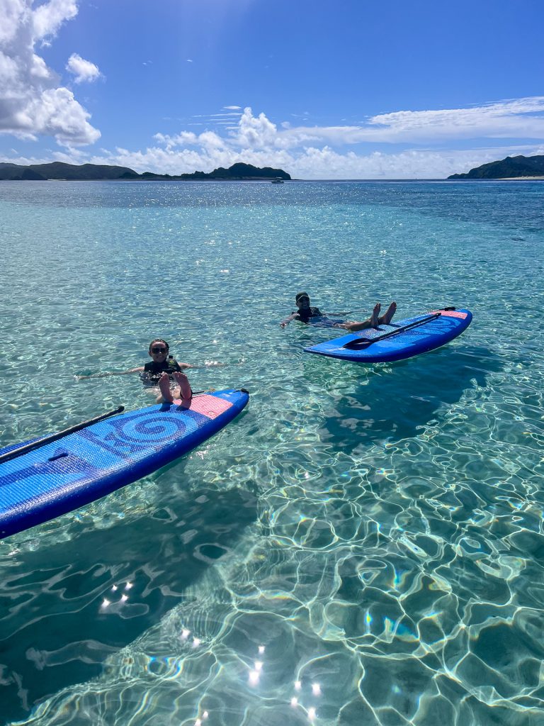 Two people floating in crystal clear waters with their feet up on paddleboards with land in the background and bright blue skies.