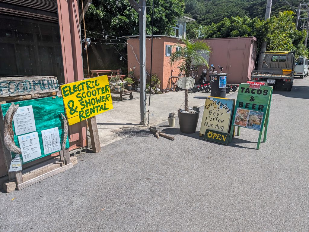 Wooden, handmade signs on the side of a road advertising scooter rental, shower rental, food, and drink for sale.