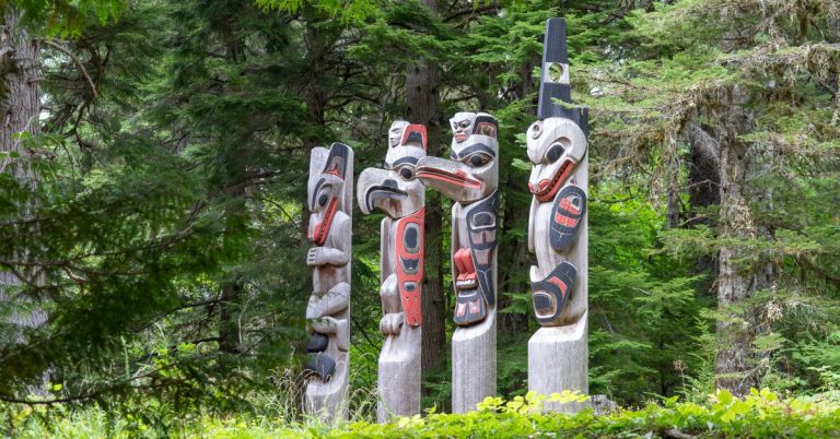 Four wooden totem poles carved and painted to look like birds and animals surrounded by pine trees.