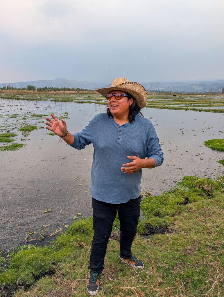 A man in a hat and blue shirt standing on grass in front of wetlands gesturing with his hand while talking.