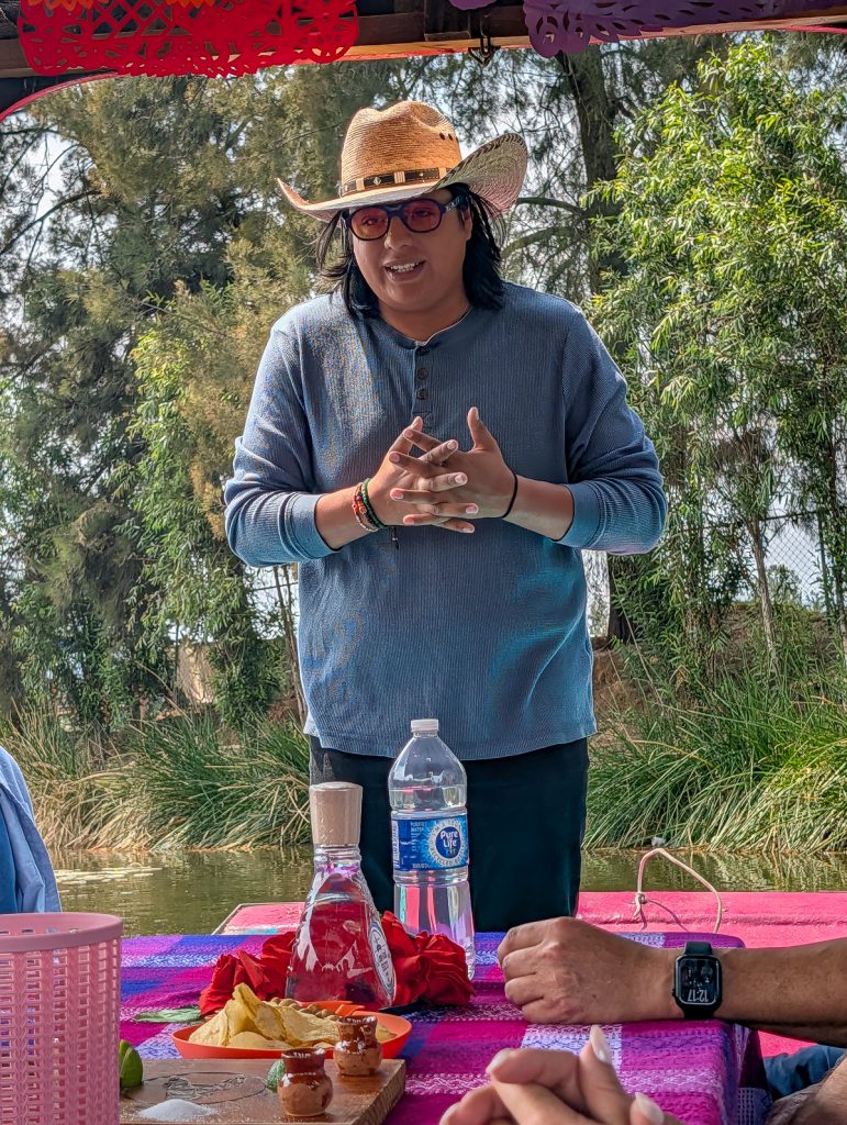 A man in a long-sleeved blue top, hat and sunglasses talking to people sitting in front of him out of shot.