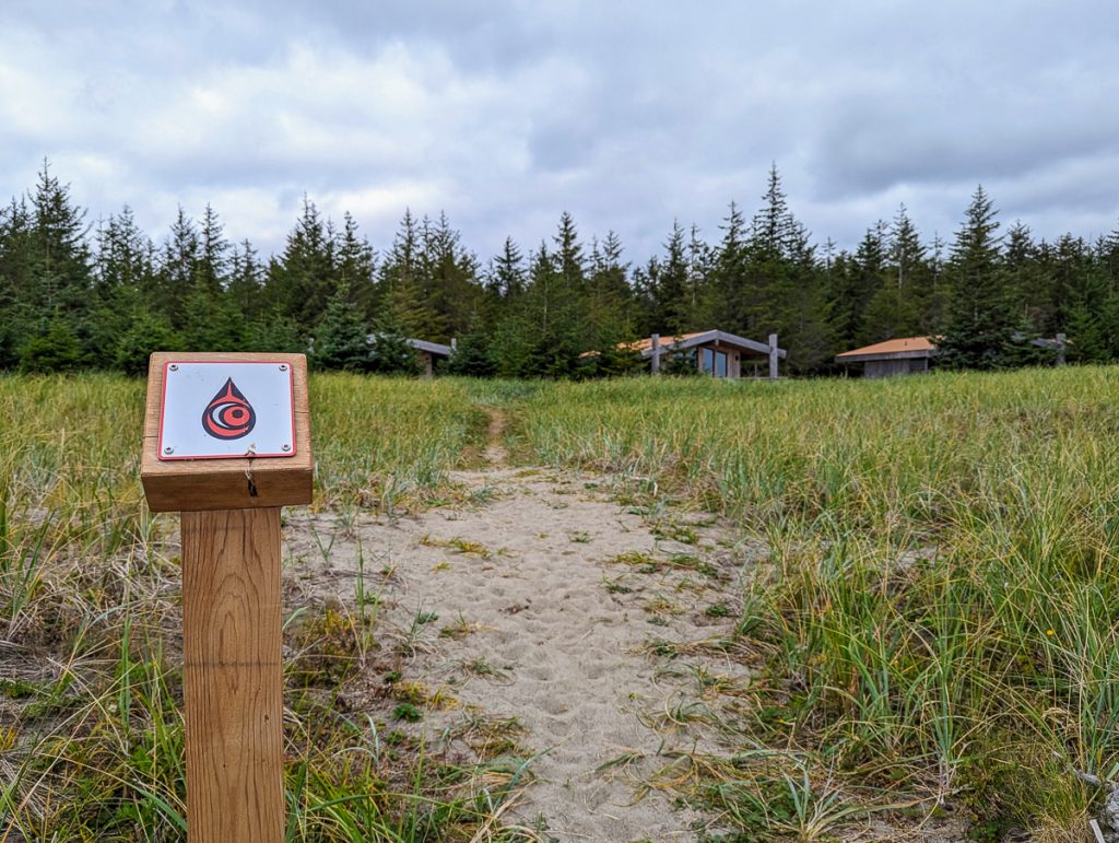 A wooden sign with a teardrop shaped logo next to a sandy path through sedge towards some wooden cabins partially hidden by pine trees.