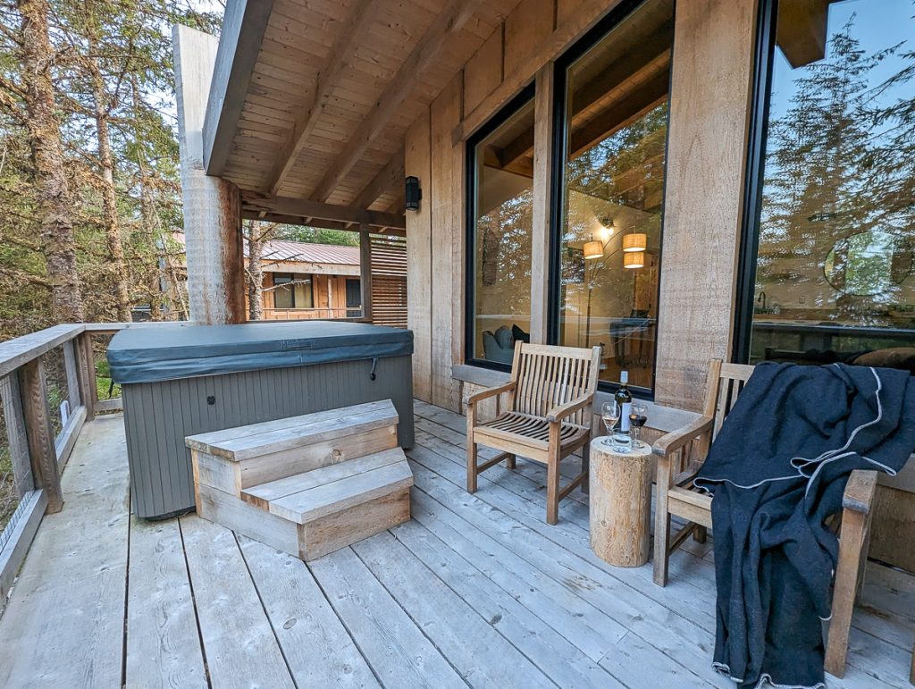A wooden deck with a hot tub with wooden steps next to it, wooden chairs and a small log being used as a table for a wine bottle and glasses.