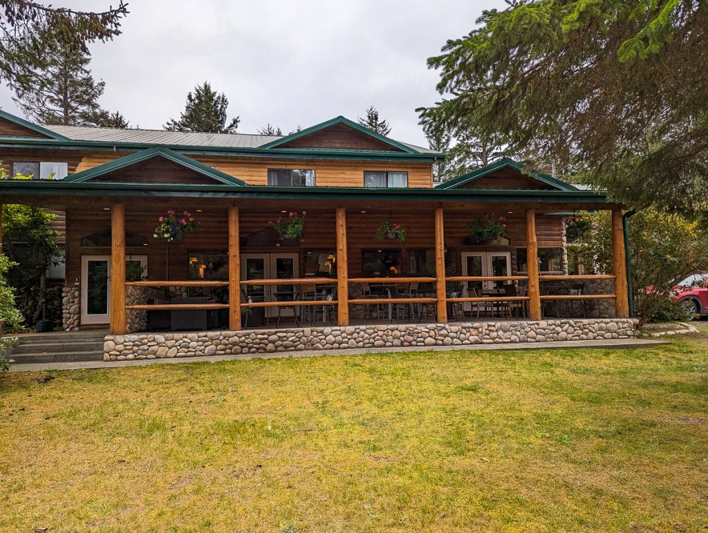 A wooden lodge with a deck and stone foundations with grass in front and some trees and gray clouds behind.