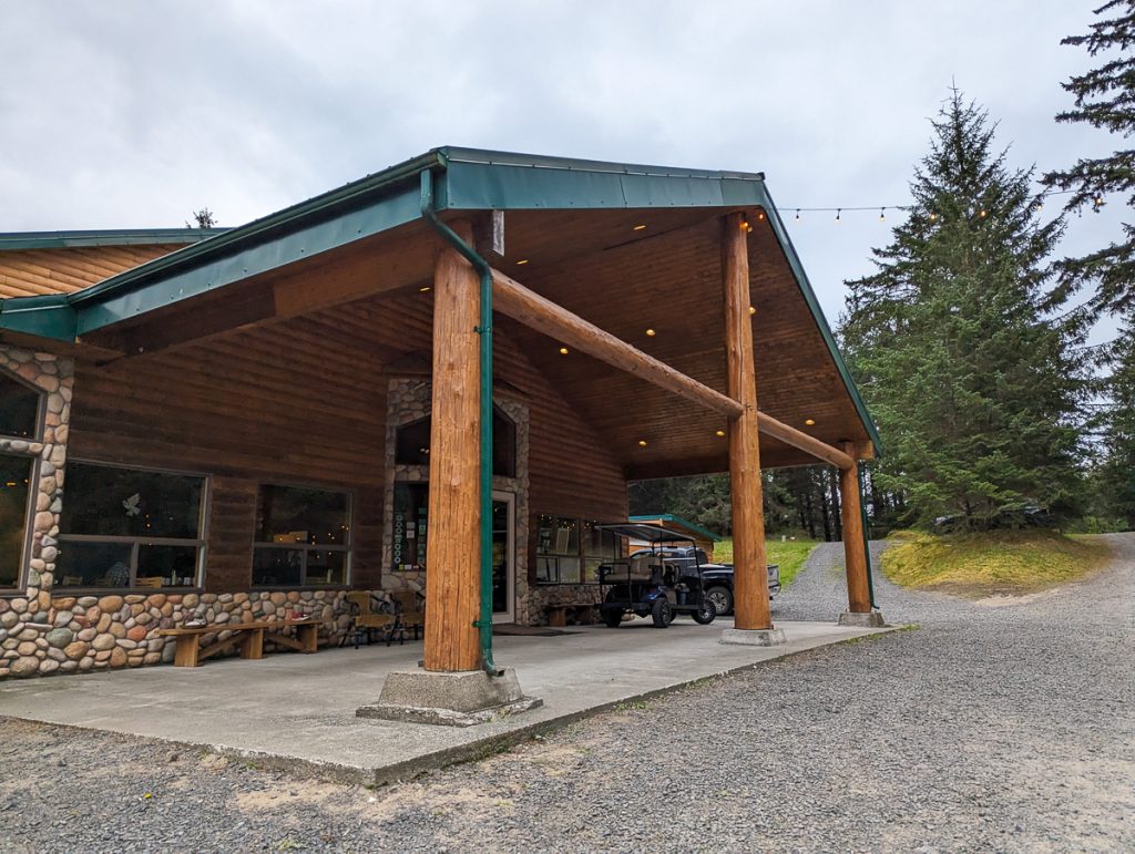 The entrance of a big wooden lodge with stone foundations and a big deck area with a golf buggy parked on concrete underneath.