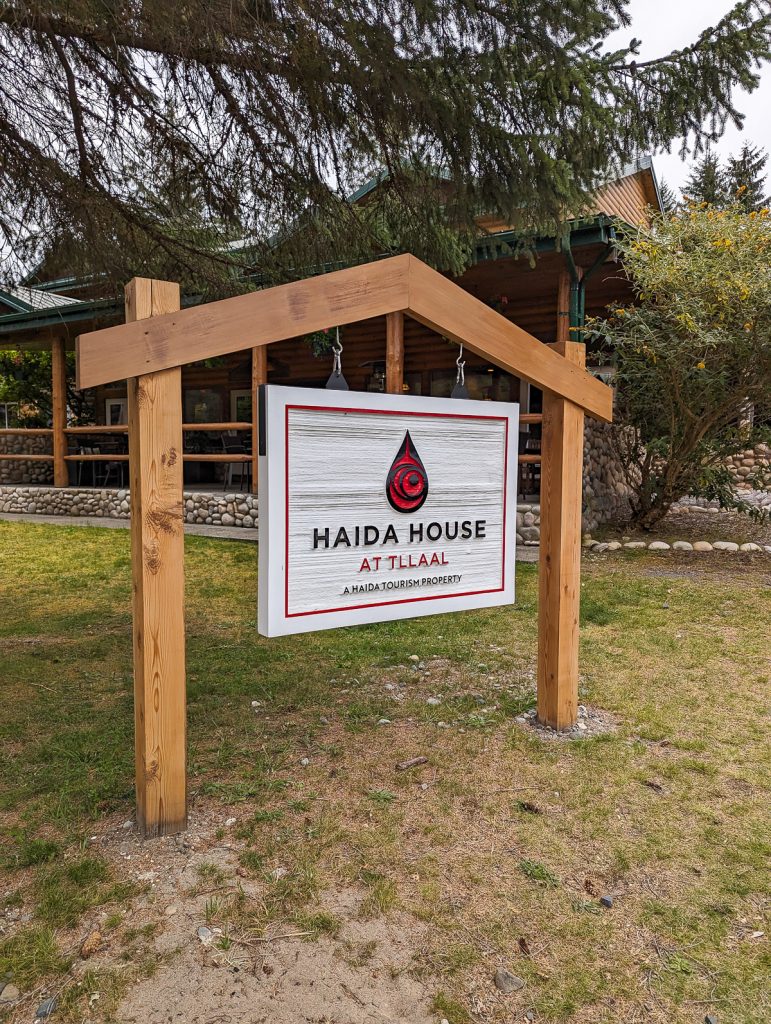 Wooden posts making the shape of a house with a white sign hanging from the "roof" that shows a black and red teardrop logo and the words "Haida House at Tllaal A Haida Tourism Property."