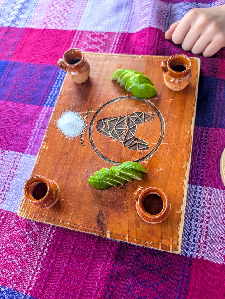 A wooden board on a patterned cloth with four small ceramic cups, slices of lime and a pile of salt.