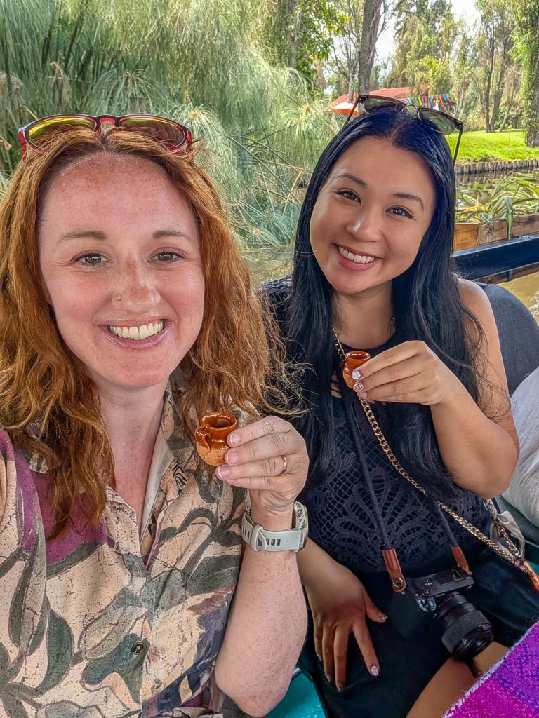 Sooz and Christina holding tiny ceramic cups and smiling at the camera with greenery behind them.