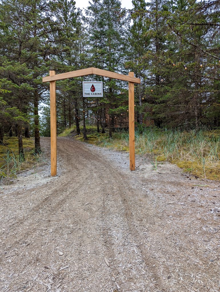 A bark pathway leading to wooden posts holding up a white sign with a red and black teardrop logo and the words "the cabins" with pine trees in the background.