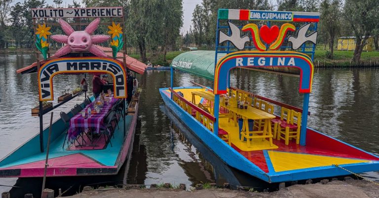 Two colorful flat bottomed boats with long tables and chairs moored on a canal.