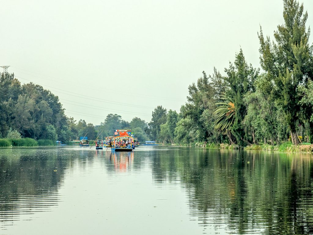 Colorful flat bottomed boats on a canal with tall trees on either side of the water. 