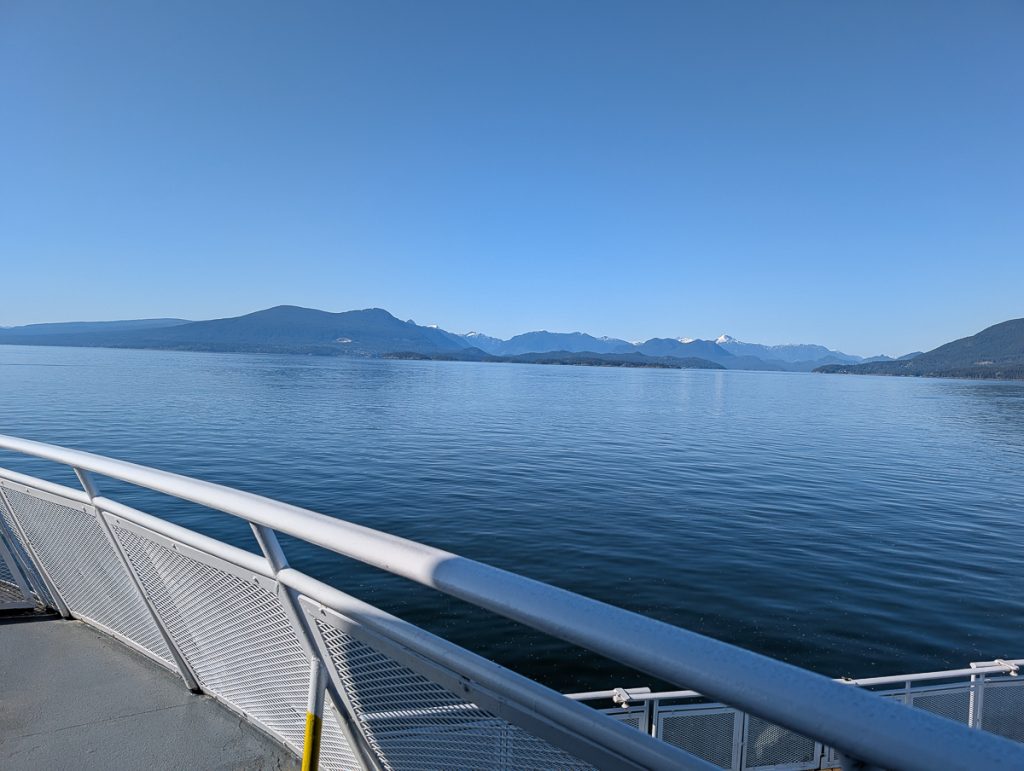 The view from a boat deck across calm blue waters towards mountains in the distance, under a clear blue sky.