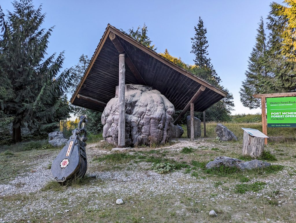 A massive wooden burl underneath a roof with pine trees behind it.