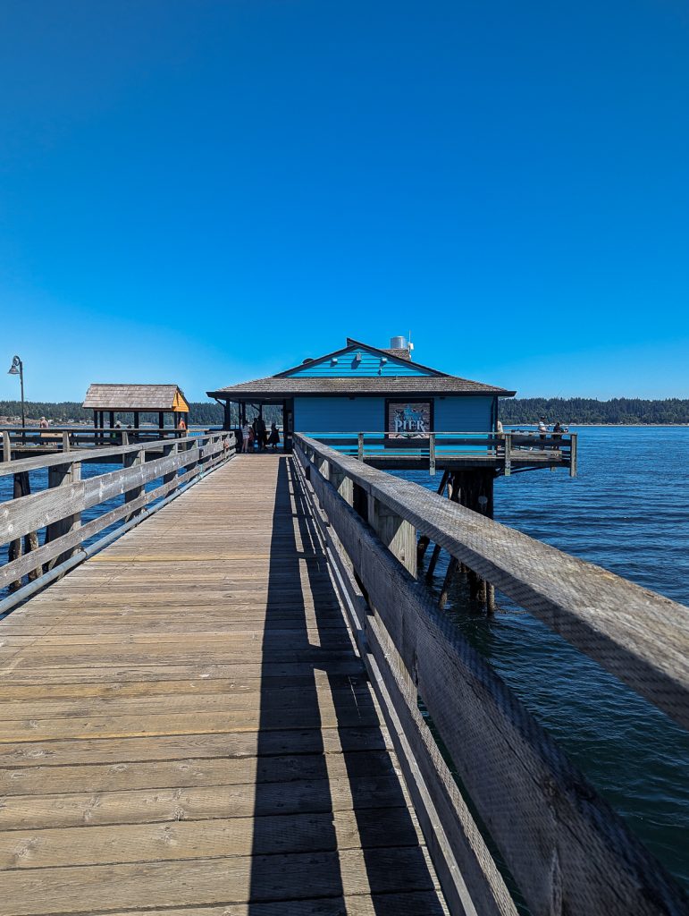 A wooden pier leading to a blue wooden building at the end looking out over dark blue water and blue skies.