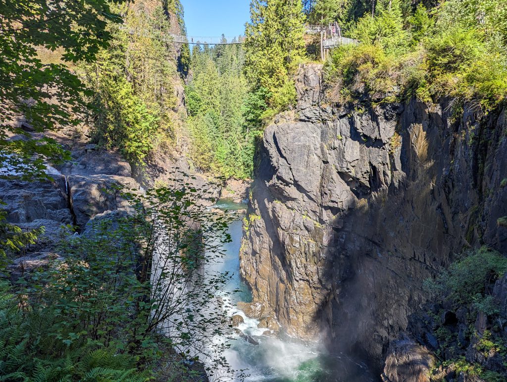 A gorge surrounded by pine trees with a waterfall at the bottom and suspension bridge at the top.
