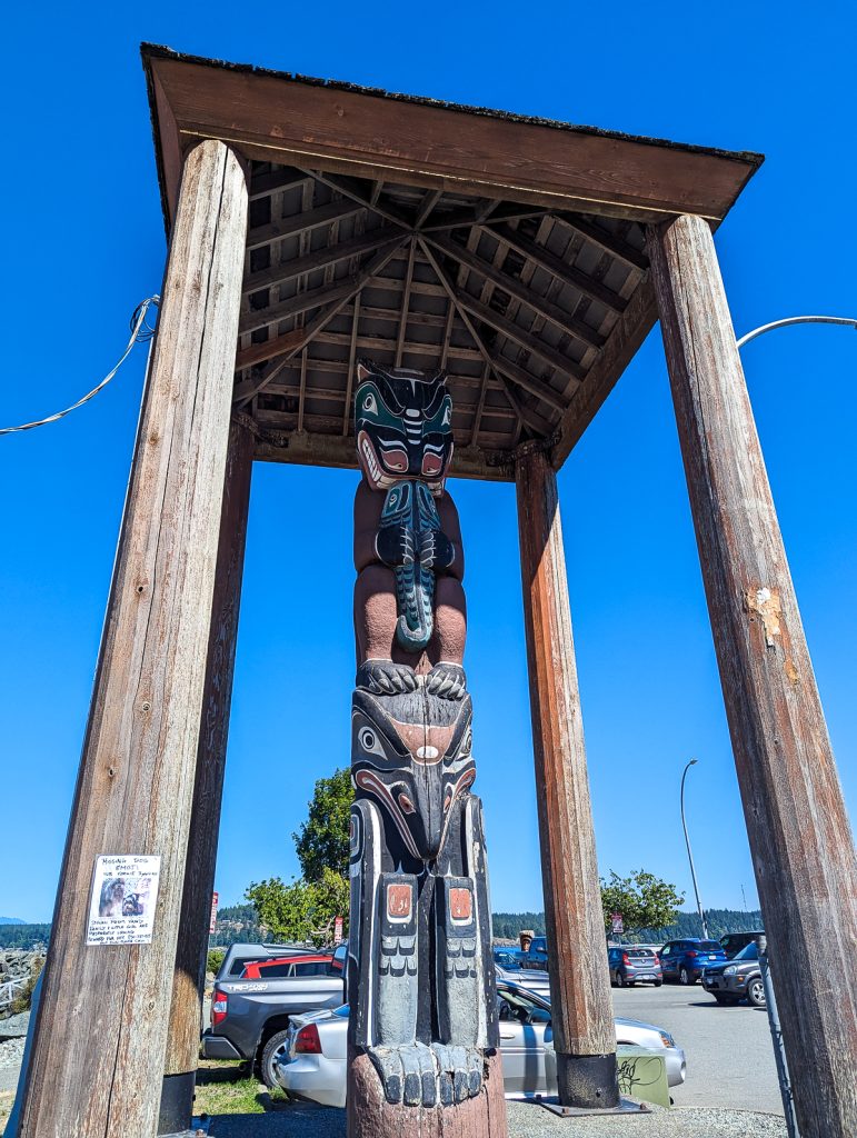 A carved and painted totem pole standing under a wooden roof on a sunny day.