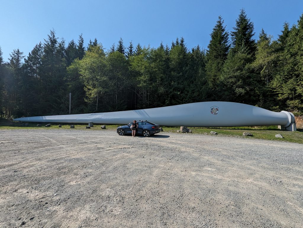 A person and a car looking tiny next to a giant wind turbine blade lying on grass next to a gravel parking lot with pine trees and blue sky behind it.