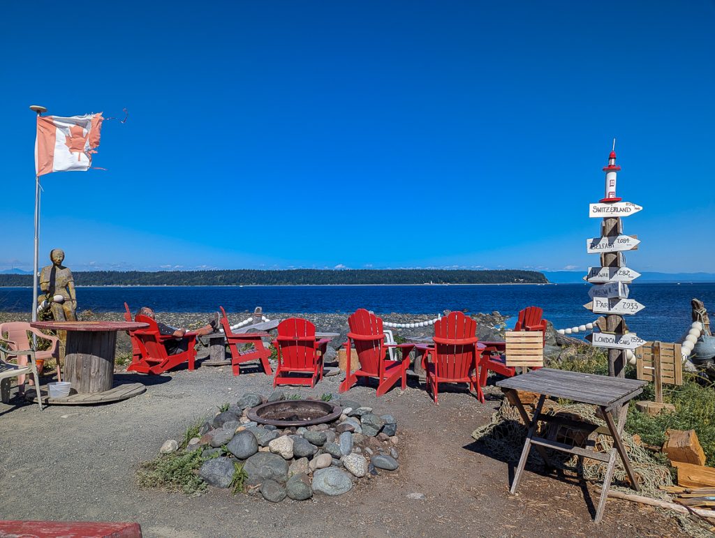A group of red wooden chairs and tables with a tattered Canadian flag and a wooden signpost all looking out over water with a promontory covered in pine trees and blue skies in the distance.