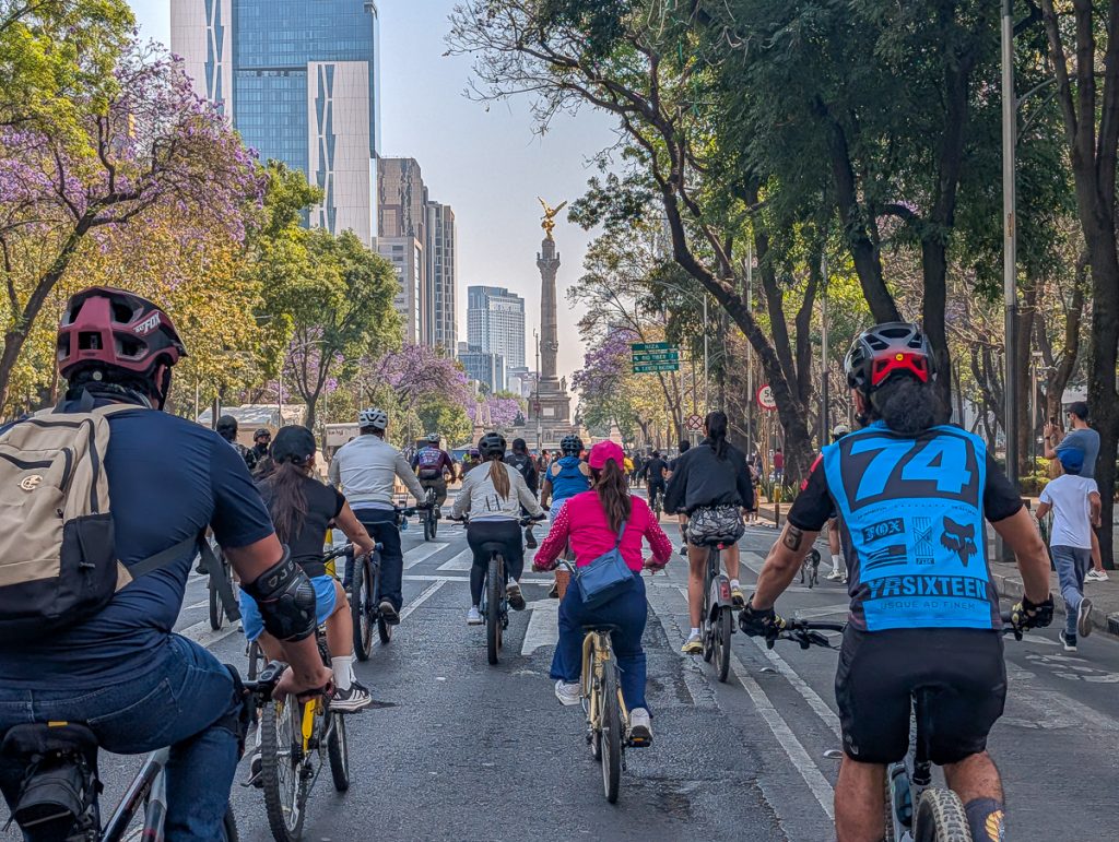A group of people ride in the middle of a road with a statue in the distance as part of Mexico City car-free Sunday