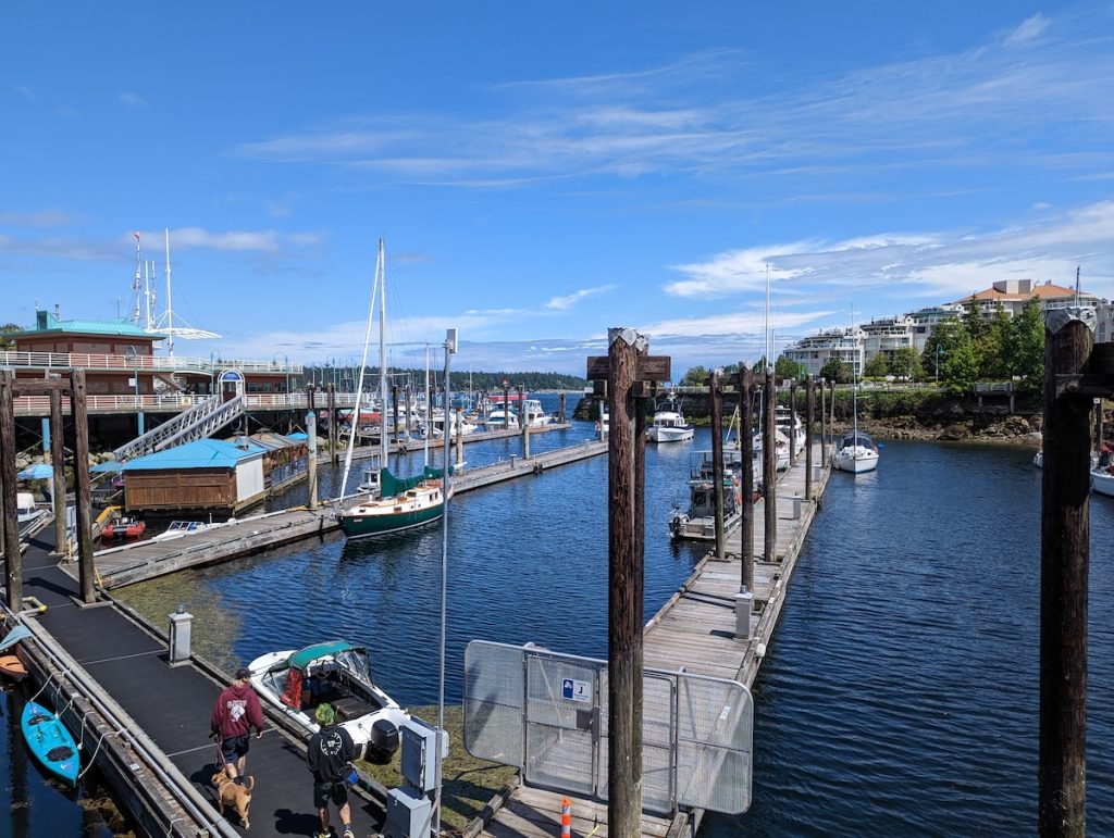 A view looking down over a marina with wooden jetties and boats moored, with buildings on land in the distance and blue skies above.