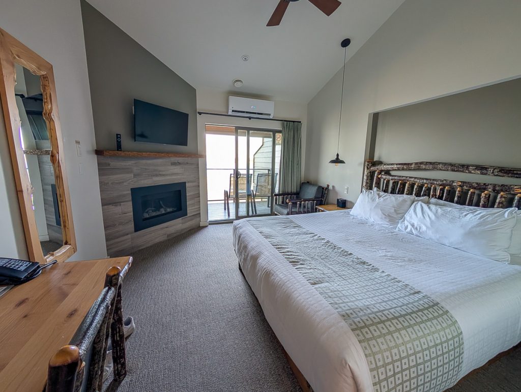 A big hotel room with a bed, desk, fireplace built into the wall and tv mounted above it.